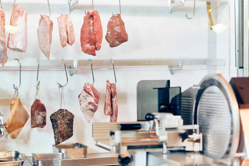Fleisch und Schinken an Haken vor weißen Fliesen, daneben eine Aufschnittmaschine
